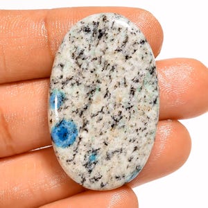 May include: An oval-shaped polished gemstone featuring a speckled white and light gray base with prominent black inclusions and distinct, vibrant blue circular patches distributed across the surface, showcasing a smooth, reflective finish.