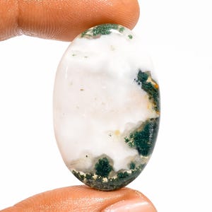 Pode incluir: Uma pedra preciosa em formato de cabochão oval com uma superfície lisa e polida sobre uma base branca leitosa. A pedra exibe inclusões dendríticas verde-escuras semelhantes a musgo, concentradas principalmente ao longo das bordas inferior e direita.
