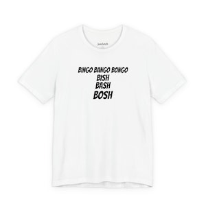 Puede incluir: Camiseta blanca con la frase "BINGO BANGO BONGO BISH BASH BOSH" en negro, centrada en la parte delantera. La camiseta tiene mangas cortas y cuello redondo. La marca "jusclutch" está impresa en la parte superior de la espalda.