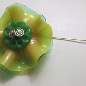 May include: A handcrafted flower ornament made from translucent green and yellow plastic material. The center features a small, circular green plastic component with a scalloped edge, accented by two silver-toned metal wire spirals and a thin wire stem.