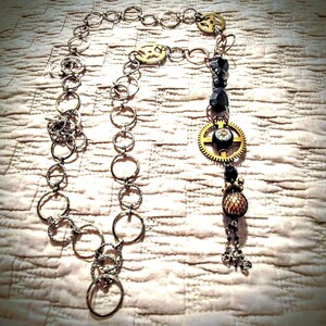 May include: A steampunk necklace with a silver chain and various ring designs. It features gold-coloured gears, black beads, and a pendant with a crystal accent. The design creates a unique, industrial aesthetic, suitable for alternative fashion.