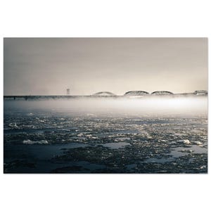 May include: A black and white photograph of a bridge over a body of water. The bridge is partially obscured by fog or mist. The water is covered in ice. The sky is overcast.