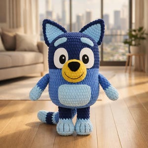 May include: A handmade crochet Bluey plush toy. The toy is predominantly blue and light blue, with a yellow nose and large eyes. It stands on a wooden floor, with a blurred background of a living room and city skyline.