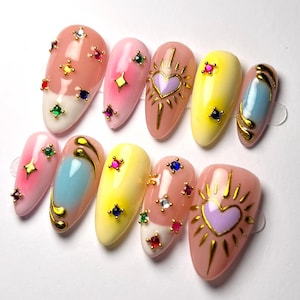 May include: A set of ten almond-shaped artificial nails. The nails feature a variety of designs, including ombre effects, gold accents, and jewel embellishments. Colors include pink, yellow, blue, and white. Some nails have heart and star designs.