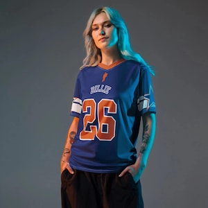 Könnte beinhalten: Ein blaues Trikot mit orangefarbenen Akzenten, das die Nummer "26" und den Namen "Billie" zeigt. Das Trikot hat weiße Streifen auf den Ärmeln und einen V-Ausschnitt. Das Trikot wird von einer Person mit blonden Haaren getragen.