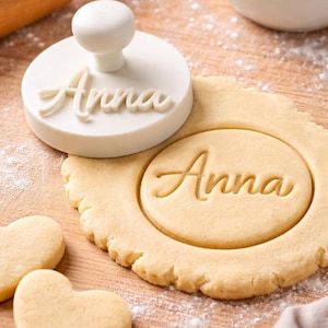 May include: A white cookie stamp with the name "Anna" is next to a round cookie with the same name imprinted. Two heart-shaped cookies are nearby. A rolling pin and a bowl of flour are in the background, all on a wooden surface.