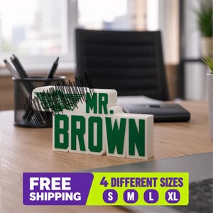 May include: A white 3D-printed desk sign with green lettering that reads MR. BROWN, featuring a green winged logo on the left side. The sign is positioned on a wooden surface, with a black pen holder and office supplies visible in the background.