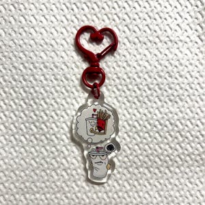 May include: A clear acrylic keychain featuring a cartoon milkshake character with a thought bubble showing a milkshake and fries embracing. The keychain has a red heart-shaped clasp.