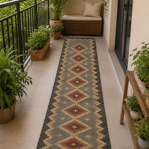 Puede incluir: Una alfombra larga y rectangular con un patrón geométrico de diamantes y zigzags. La alfombra presenta un fondo gris con formas de diamantes rojos, beige y negros. La alfombra está colocada sobre una superficie embaldosada y tiene un borde con flecos.