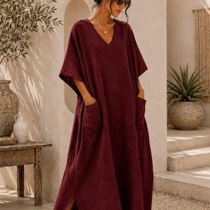 May include: A deep burgundy, loose-fitting maxi dress featuring a V-neckline, short wide sleeves, and two large patch pockets on the front. The garment is crafted from a textured fabric and includes side slits at the hem for ease of movement.