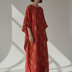 May include: A red, long-sleeved dress with a yellow animal print. The dress has a loose fit and a round neckline. The model is wearing white trainers. The text "Trend Trunk Syndicate" is at the bottom of the image.