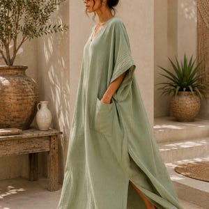 May include: A sage green maxi dress with a V-neckline, short wide sleeves, and a large patch pocket on the side. The garment features a textured fabric, a loose silhouette, and a high side slit. The text TrendTrunkSyndicate is visible.