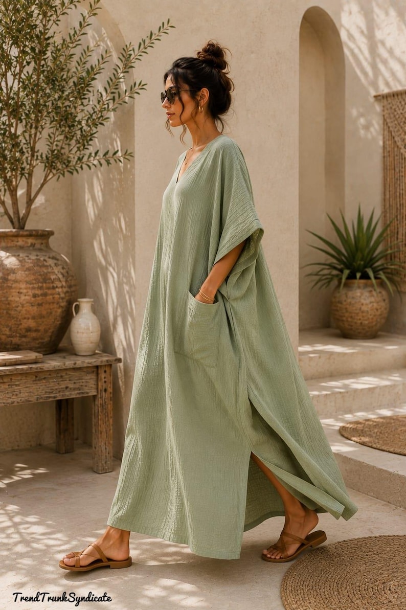 May include: A sage green maxi dress with a V-neckline, short wide sleeves, and a large patch pocket on the side. The garment features a textured fabric, a loose silhouette, and a high side slit. The text TrendTrunkSyndicate is visible.