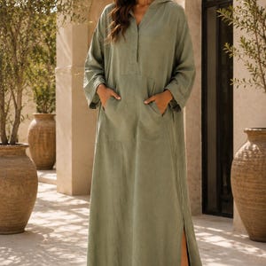 May include: A floor-length sage green hooded dress with long sleeves, a V-neckline, and a large front kangaroo pocket. The garment features a loose, relaxed fit with side slits at the hem and is crafted from a textured fabric. TrendTrunkSyndicate.
