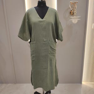 May include: A sage green midi dress featuring a V-neckline, short wide sleeves, and two large patch pockets on the front. The garment is displayed on a black mannequin stand and is crafted from a textured, lightweight fabric with side slits.