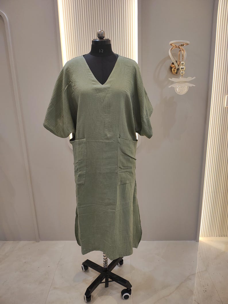 May include: A sage green midi dress featuring a V-neckline, short wide sleeves, and two large patch pockets on the front. The garment is displayed on a black mannequin stand and is crafted from a textured, lightweight fabric with side slits.