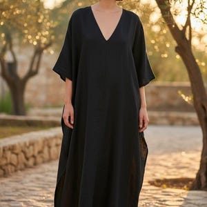 May include: A black maxi dress with a V-neck and short sleeves. The dress is made of a flowing fabric and is worn outdoors. The image is taken in natural light, with trees and a stone wall in the background.