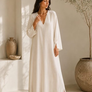 May include: A floor-length, off-white kaftan dress featuring long, wide sleeves with split cuffs, a deep V-neckline, and side slits at the hem. The garment is paired with simple, closed-toe white slip-on shoes. The text TrendTrunkSyndicate is visible.