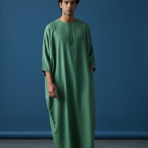 Op de afbeelding: Een lange, vloeiende groene jurk met een geknoopte halslijn en zakken. De loszittende jurk heeft driekwart mouwen en valt tot op de enkels. Het kledingstuk is gemaakt van een zachte, draperende stof. De tekst "Trend Trunk Syndicate" staat onderaan.