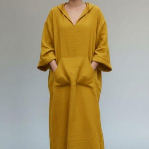 Puede incluir: Vestido con capucha amarillo mostaza con bolsillo delantero y mangas largas. El vestido tiene un corte holgado y un escote en V. La modelo lleva el vestido con las manos en los bolsillos. El texto "TRENDTRUNKSYNDICATE" está en la parte inferior.