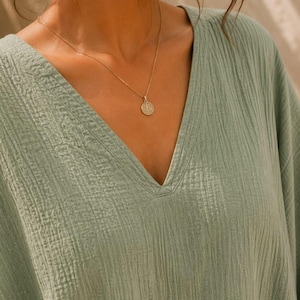 May include: A sage green V-neck top made of textured, crinkled fabric. The garment features a simple neckline and is paired with a delicate gold chain necklace featuring a small, round, embossed pendant. The text TrendTrunkSyndicate is visible.