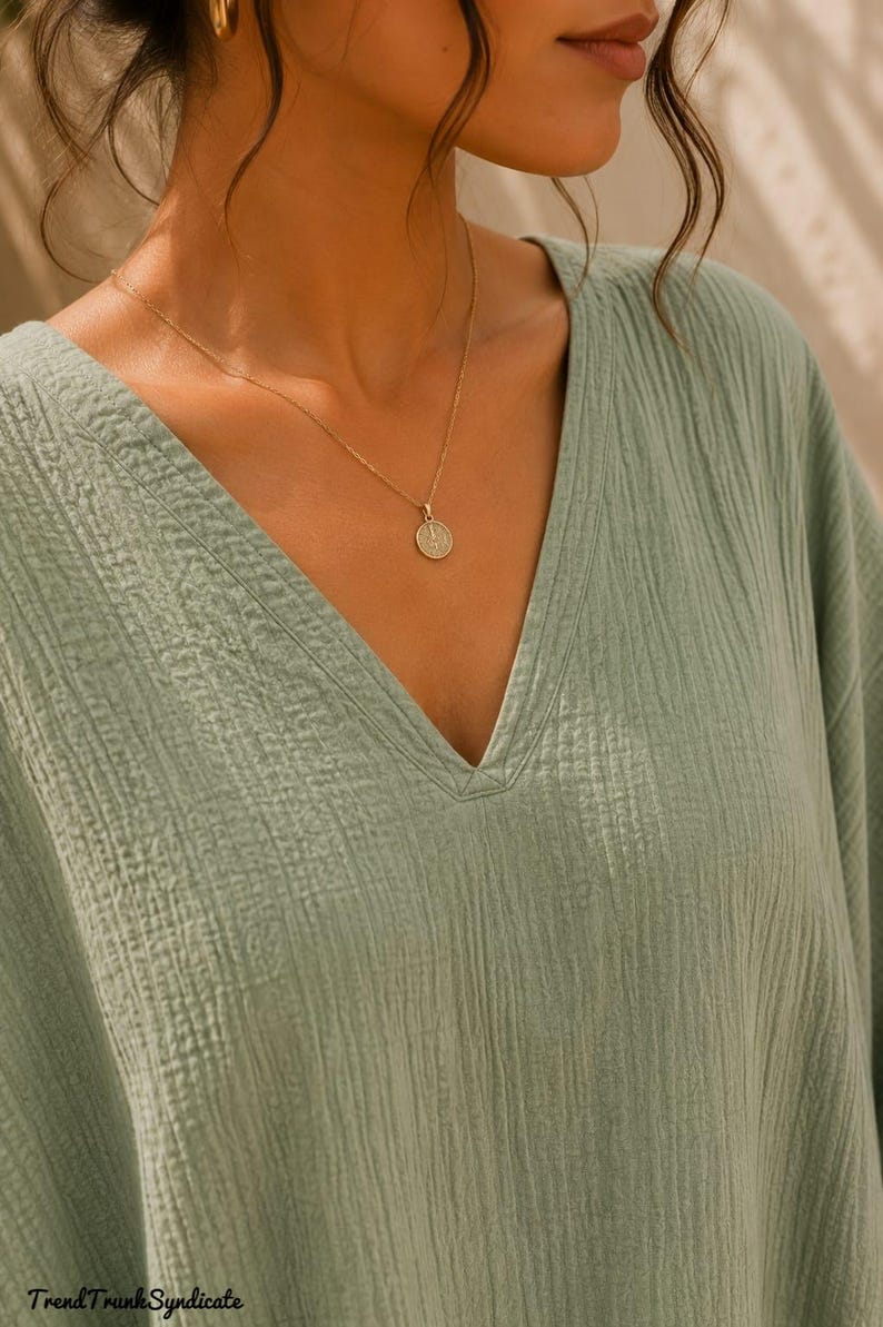 May include: A sage green V-neck top made of textured, crinkled fabric. The garment features a simple neckline and is paired with a delicate gold chain necklace featuring a small, round, embossed pendant. The text TrendTrunkSyndicate is visible.