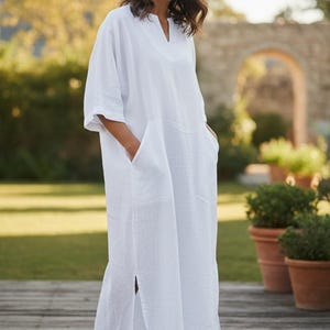 May include: A white linen dress with a V-neck and short sleeves. The dress has two large pockets and a slit on the side. The model is standing on a wooden deck in front of a garden.