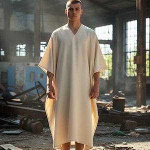 May include: A cream-coloured, V-neck caftan with wide sleeves. The garment is worn by a person standing barefoot in a dilapidated industrial space. The caftan's fabric drapes loosely, creating a flowing silhouette.