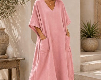 May include: A loose-fitting, ankle-length dress in a soft pink hue, featuring a V-neckline, short wide sleeves, and two large patch pockets on the front. The garment has a textured fabric finish and includes side slits at the hem for movement.