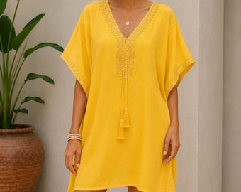 May include: A bright yellow V-neck tunic dress featuring short kimono-style sleeves, intricate embroidery along the neckline, and two hanging tassels. The garment has a loose, relaxed fit and includes subtle decorative trim along the sleeve edges.