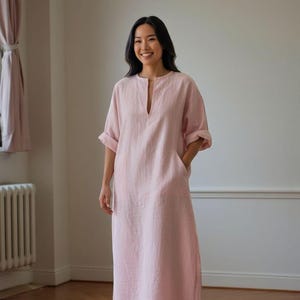 May include: A light pink linen dress with a V-neckline and short sleeves. The dress has two front pockets and a relaxed fit. The model is standing in a room with wooden floors and a radiator.