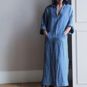 Puede incluir: Una larga túnica azul con capucha, cuello en V y bolsillos delanteros. La túnica está hecha de una tela suave y fluida y tiene mangas largas. La persona lleva zapatos negros. El texto "TRENDTRUNKSYNDICATE" está en la parte inferior.