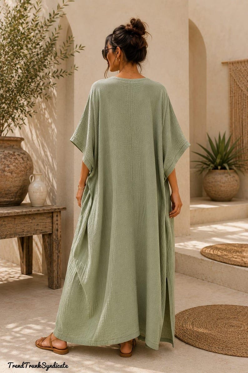 May include: A sage green maxi kaftan dress with a textured, crinkled fabric finish, wide short sleeves, and side slits. The garment features a loose, flowing silhouette and a simple round neckline. The text TrendTrunkSyndicate is visible.