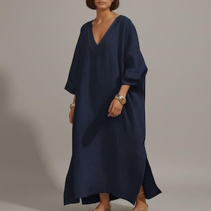 Peut inclure: Longue robe bleu marine à col en V avec des manches larges. La robe a des fentes latérales et est portée avec des bracelets dorés et des sandales. Le texte "Trend Trunk Syndicate" est au bas de l'image.