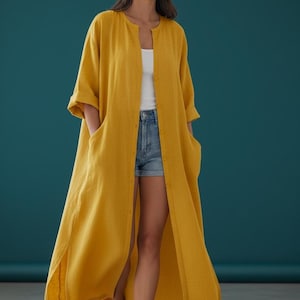 May include: A long, mustard yellow linen duster coat with large pockets and a button-down front. The coat is worn over a white top and denim shorts. The image is set against a teal background. The brand name "Trend Trunk Syndicate" is visible at the bottom.