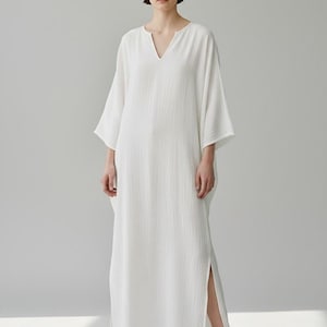 May include: A floor-length white kaftan dress featuring a textured fabric, a V-neckline, and wide three-quarter length sleeves. The garment has side slits at the hem and is paired with white sneakers. The text TrendTrunkSyndicate is visible.