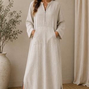 May include: A floor-length beige dress with long sleeves, a front zip closure at the neckline, and a large kangaroo-style pocket on the front. The garment features side slits at the hem and is made of a textured fabric. The text TrendTrunkSyndicate is visible.