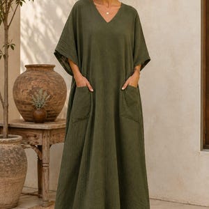 May include: An olive green maxi dress featuring a V-neckline, short wide sleeves, and two large front patch pockets. The garment is crafted from a textured fabric and falls to the ankles. The text TrendTrunkSyndicate is visible in the bottom corner.