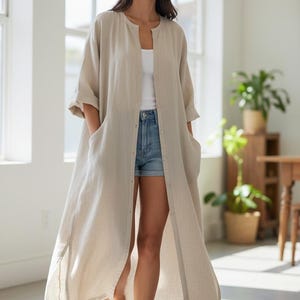 May include: A long, beige linen duster coat with a button-up front and pockets. The coat is worn over a white top and denim shorts. The image is taken indoors with natural light.