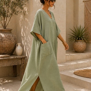 May include: A sage green V-neck maxi dress with short wide sleeves, two large front patch pockets, and side slits at the hem. The garment features a textured fabric finish and is paired with brown flat sandals. The text TrendTrunkSyndicate is visible.