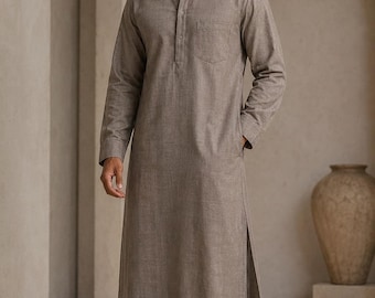 May include: A long-sleeved, ankle-length taupe tunic featuring a mandarin collar, a partial button-down placket, and a single chest pocket. The garment has side slits at the hem and is paired with white sneakers. The text TrendTrunkSyndicate is visible.