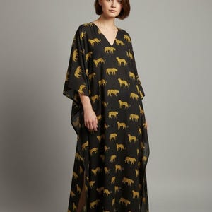 May include: Black, long, V-neck dress with a gold leopard print. The dress has wide sleeves and a slit on the side. The model is wearing white sneakers. The dress is a maxi length.