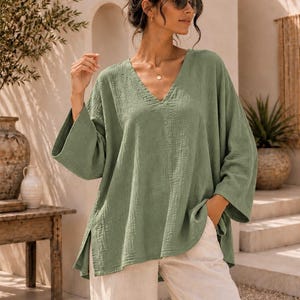 May include: A sage green V-neck tunic top with a textured, crinkled fabric finish, wide three-quarter length sleeves, and side slits at the hem. The garment is paired with loose-fitting, off-white trousers. The text TrendTrunkSyndicate is visible.