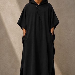 May include: A floor-length black hooded kaftan featuring short, wide sleeves, a single button closure at the neckline, and side slits. The garment has a loose, flowing silhouette and includes deep side pockets. The text TrendTrunkSyndicate is visible.