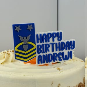 Può includere: Una torta di compleanno con glassa bianca e una decorazione. La decorazione presenta un emblema militare blu e giallo e le parole "HAPPY BIRTHDAY ANDREW!" in blu. La torta ha una base marrone chiaro.