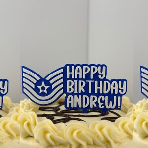 Peut inclure: Un gâteau d'anniversaire décoré avec du glaçage blanc et un décor de gâteau bleu et blanc avec l'inscription "HAPPY BIRTHDAY ANDREW!". Le décor a un design de style militaire.