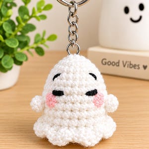 May include: A white crocheted ghost keychain featuring black yarn eyes and pink yarn cheeks. The ghost is attached to a silver-toned metal split ring by a short chain. The crochet work is textured with visible stitches throughout the entire figure.