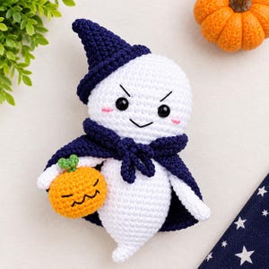 May include: A white crocheted ghost plush toy wearing a dark blue pointed hat and a matching cape. The ghost has black eyes, a stitched mouth, and pink cheeks. It holds a small orange crocheted pumpkin with a green stem and a stitched face.