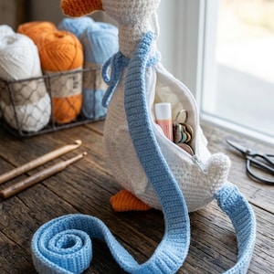Op de afbeelding: Een handgemaakte witte gehaakte gansvormige tas met een oranje snavel en poten en een blauwe band. De tas is open en onthult een rits en kleine items binnenin. Haaknaalden en garen staan op de achtergrond.