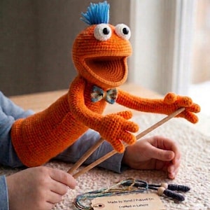 May include: An orange crocheted puppet with blue hair, large white eyes, and a bow tie. The puppet is being manipulated with wooden sticks. The puppet has a wide open mouth and is made by Hand / Puppet Co. Crafted in Lahore.